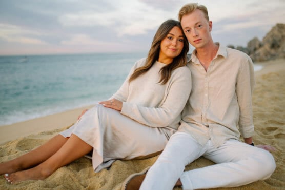 A couple sits close together on a sandy beach at sunset, both wearing light-colored clothing. The ocean and rocky cliffs are visible in the background, with a soft, cloudy sky above. by Justin Salem Meyer