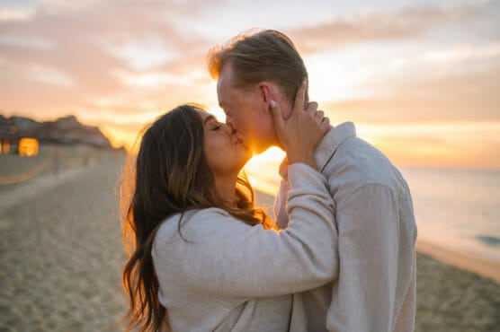 A couple shares a kiss on a beach at sunset. The sun shines behind them, creating a warm, glowing light. The woman has long dark hair and both wear light-colored sweaters. The ocean and sand are visible in the background. by Justin Salem Meyer