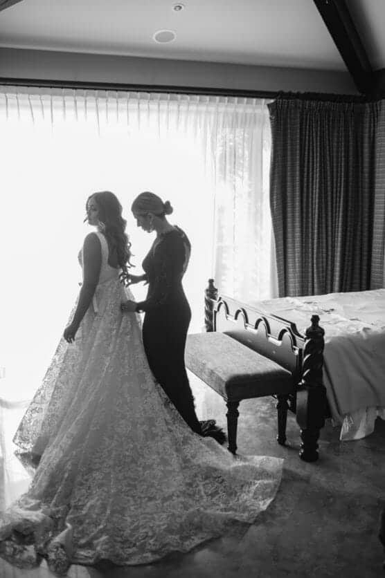 A woman in a bridal gown stands near a window as another woman helps adjust her dress. Sunlight streams in through sheer curtains, creating a soft and intimate scene in a bedroom. by Justin Salem Meyer