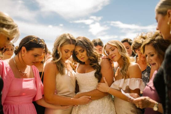 A bride in a white dress stands surrounded by supportive women, some with eyes closed, holding hands and embracing her under a bright blue sky. The mood appears emotional and heartfelt. by Justin Salem Meyer