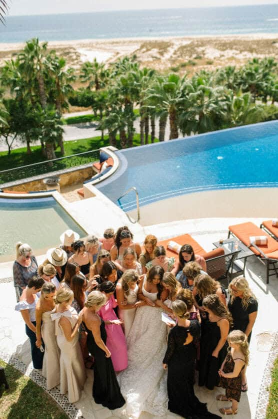 A group of women gather closely around a bride in a white dress near a pool with lounge chairs, palm trees, and the ocean visible in the background on a sunny day. by Justin Salem Meyer