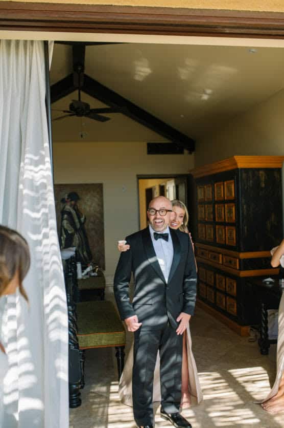 A smiling man in a black tuxedo stands indoors, with a woman in a light dress hugging him from behind. Sunlight streams through blinds, casting shadows. Another person in a light dress is partially visible on the left. by Justin Salem Meyer