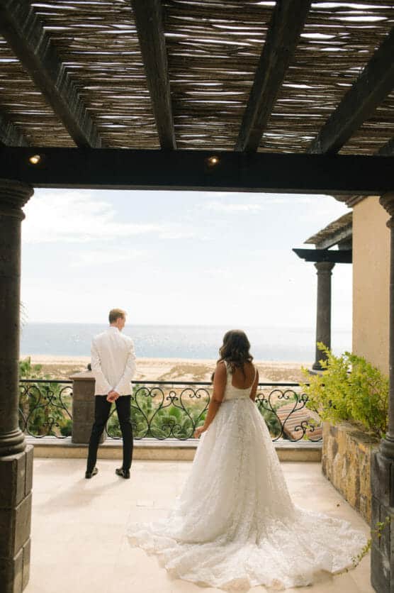 A bride in a white gown approaches a groom, who stands with his back turned, on a terrace overlooking the ocean under a wooden pergola. by Justin Salem Meyer