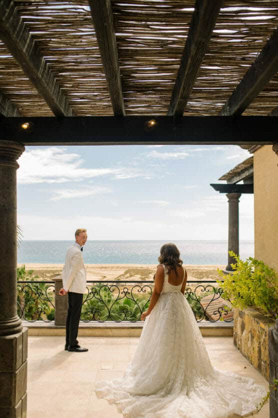 A bride in a white gown walks toward a groom in a white jacket, standing under a shaded terrace with an ocean and sandy beach visible in the background. by Justin Salem Meyer