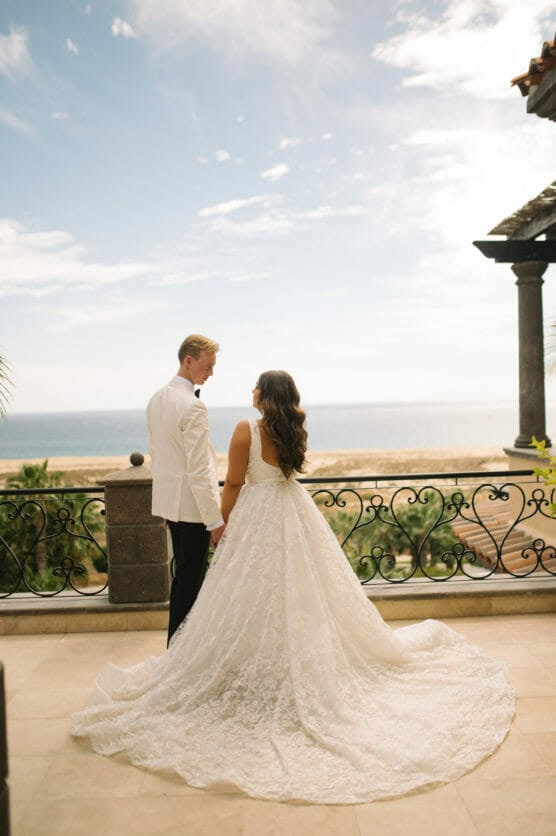 A bride in a white lace gown and a groom in a white jacket stand hand in hand on a terrace, looking out at a sunny beach and ocean under a partly cloudy sky. by Justin Salem Meyer