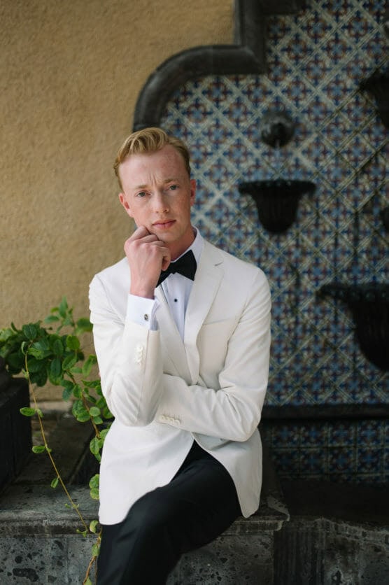 A man in a white tuxedo jacket, black bow tie, and black pants sits on a stone ledge with one hand resting on his chin. Behind him is a mosaic-tiled wall with decorative water fountains and green leaves. by Justin Salem Meyer