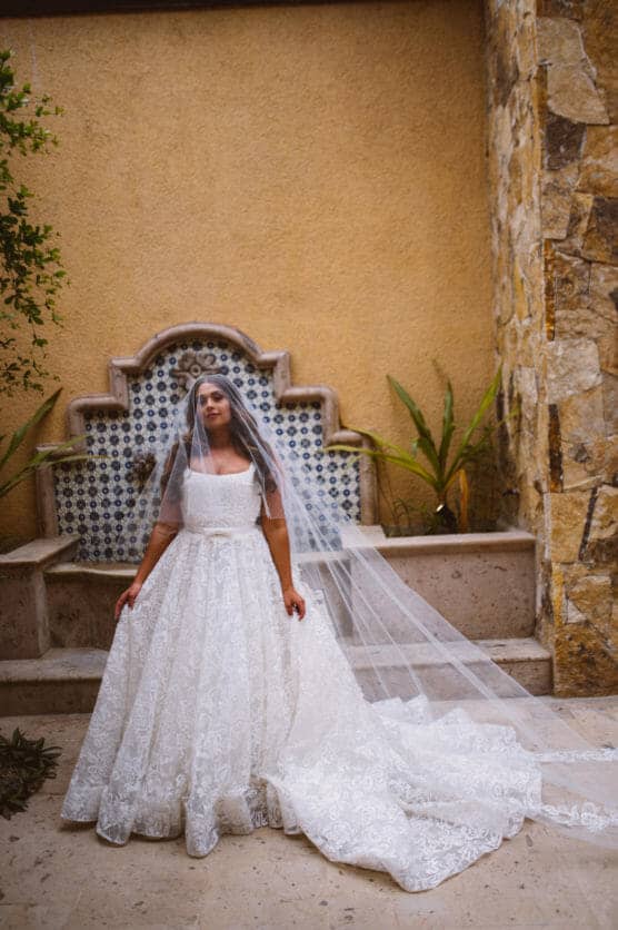 A bride in a white lace wedding gown and long veil stands outdoors in front of a decorative stone wall with patterned tiles, her eyes closed and hands resting on her dress. by Justin Salem Meyer