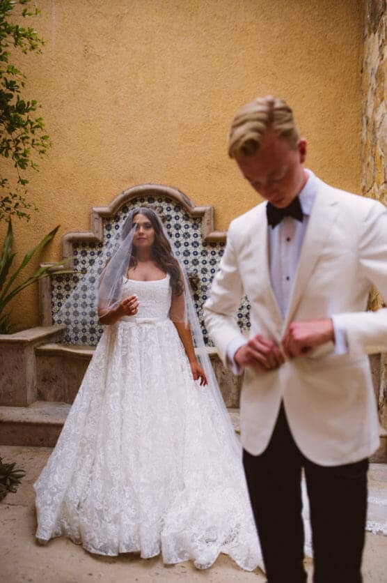 A bride in a white lace gown and veil stands in the background, while a groom in a white tuxedo jacket adjusts his jacket in the foreground. They are outdoors near a tiled wall and fountain. by Justin Salem Meyer