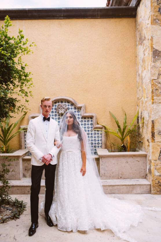 A bride in a lace wedding dress and veil stands beside a groom in a white jacket and black pants, holding hands in front of a yellow wall with a decorative tile fountain and green plants. by Justin Salem Meyer