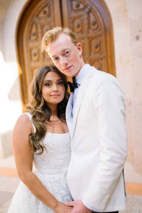 A bride and groom stand closely together in front of ornate wooden doors. The bride wears a lace white dress and the groom wears a white suit jacket with a black bow tie. They look calmly at the camera. by Justin Salem Meyer