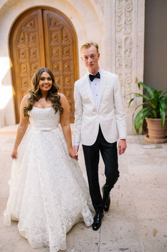 A bride in a white lace gown and a groom in a white tuxedo jacket hold hands and smile, standing in front of a large wooden arched door with ornate stonework. by Justin Salem Meyer