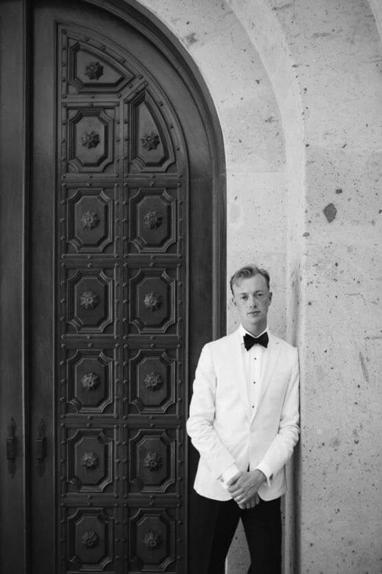 A person in a white tuxedo jacket and black bow tie stands against a stone wall next to a large, ornate wooden door with geometric patterns. The photo is in black and white. by Justin Salem Meyer