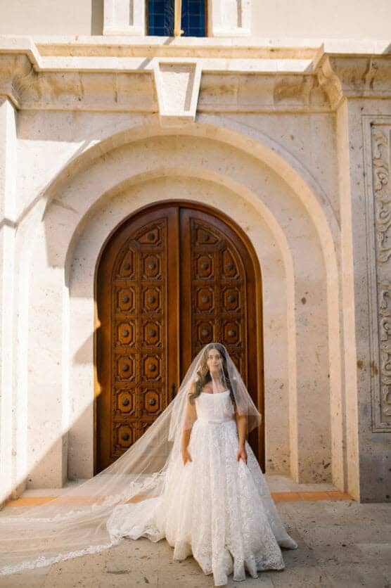 A bride in a white lace wedding gown and long veil stands in front of an ornate wooden door framed by light stone arches, with sunlight casting soft shadows around her. by Justin Salem Meyer