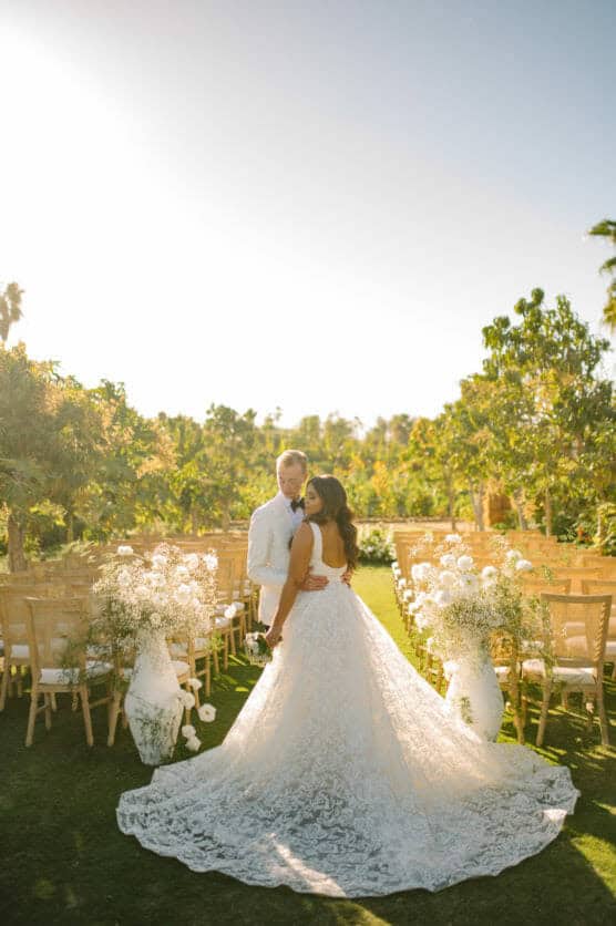 A bride in a lace wedding gown and a groom in a white suit stand close together, smiling, in an outdoor garden aisle decorated with white flowers and wooden chairs under a clear, sunny sky. by Justin Salem Meyer