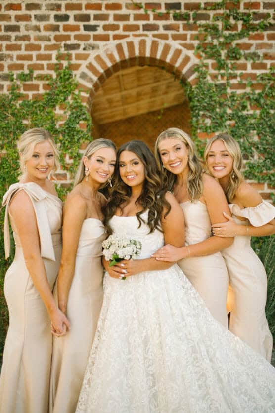 A bride in a white gown holding a bouquet stands smiling with four bridesmaids in matching beige dresses, posing together in front of a brick wall with a green vine and an arched doorway. by Justin Salem Meyer