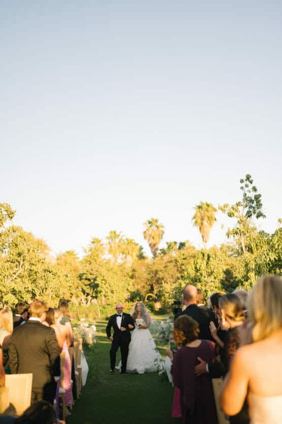 A bride in a white gown walks down an outdoor aisle with an older man, surrounded by seated and standing guests, with green trees and palm trees in the background under a clear sky. by Justin Salem Meyer