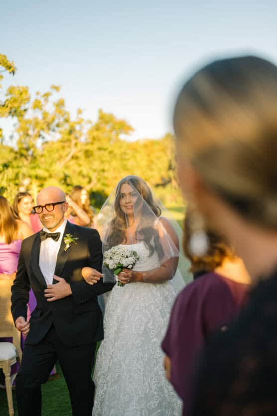 A bride in a white gown and veil, holding a bouquet, walks outside on green grass with a man in a black suit and bow tie. People in colorful dresses observe in the background under a clear sky. by Justin Salem Meyer