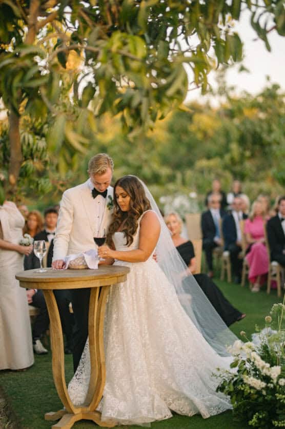 A bride in a white lace gown and a groom in a white tuxedo jacket stand together at a round wooden table outdoors, surrounded by guests seated on a lawn at a wedding ceremony. by Justin Salem Meyer