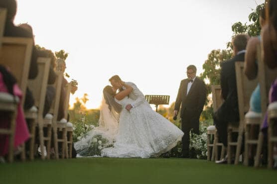 Two brides in white wedding dresses share a kiss at an outdoor ceremony, surrounded by guests seated in rows. A person in a suit stands beside them, and the sun sets in the background. by Justin Salem Meyer