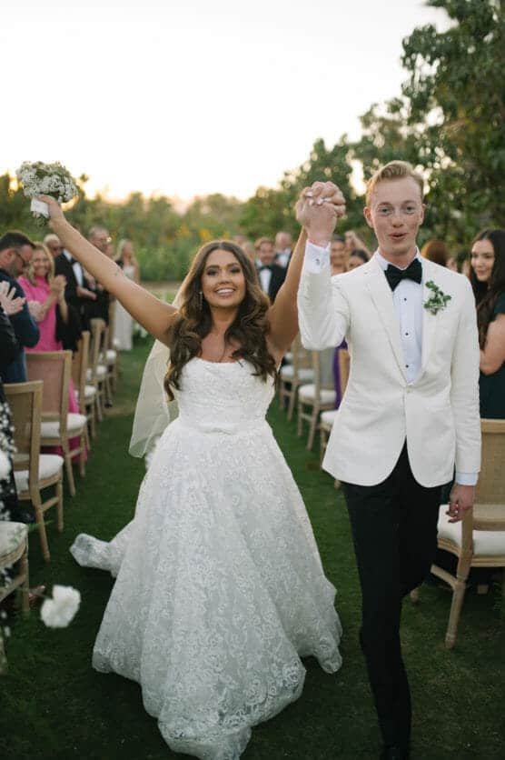 A newlywed couple walks down the aisle outdoors, holding hands and smiling joyfully. The bride wears a white lace gown and holds a bouquet; the groom wears a white jacket and black bow tie. Guests clap and celebrate in the background. by Justin Salem Meyer