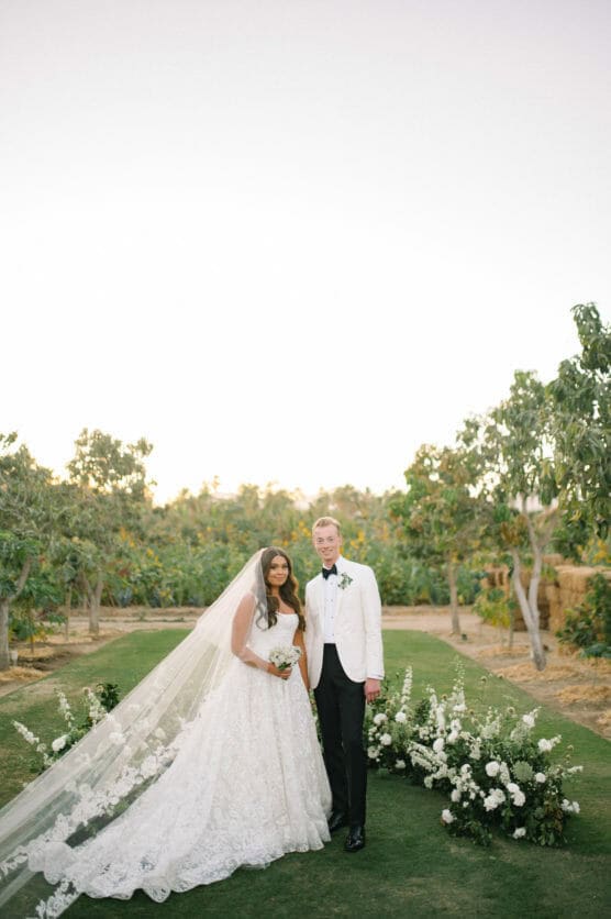A bride in a lace gown with a long veil and a groom in a white tuxedo jacket stand together outdoors on a grassy aisle, surrounded by flowers and trees, under a clear sky. by Justin Salem Meyer