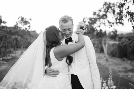 A bride and groom embrace and smile joyfully outdoors, with the bride wearing a veil and white gown and the groom in a white jacket and bow tie, surrounded by trees in a natural setting. by Justin Salem Meyer