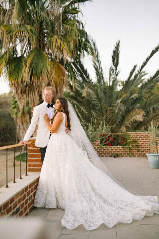 A bride in a white gown and long veil leans against a groom in a white jacket and black bow tie. They stand on a brick terrace with palm trees in the background, gazing peacefully together. by Justin Salem Meyer