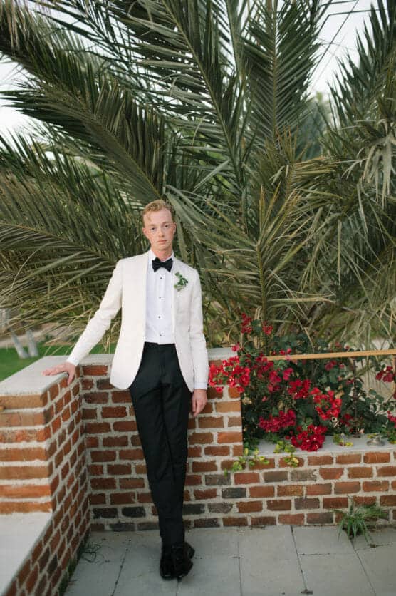 A person in a white tuxedo jacket, black bow tie, and black pants stands against a brick wall adorned with red flowers, with tall green palm leaves in the background. by Justin Salem Meyer