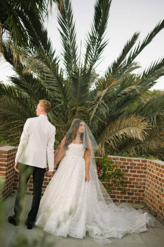 A bride in a white gown and veil holds hands with a groom in a white jacket as he faces away. They stand on a brick patio with lush palm trees in the background. by Justin Salem Meyer