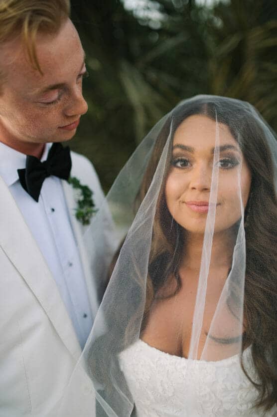 A bride in a lace dress and veil smiles at the camera while a groom in a white tuxedo jacket and black bow tie gazes at her lovingly, standing together outdoors with greenery in the background. by Justin Salem Meyer
