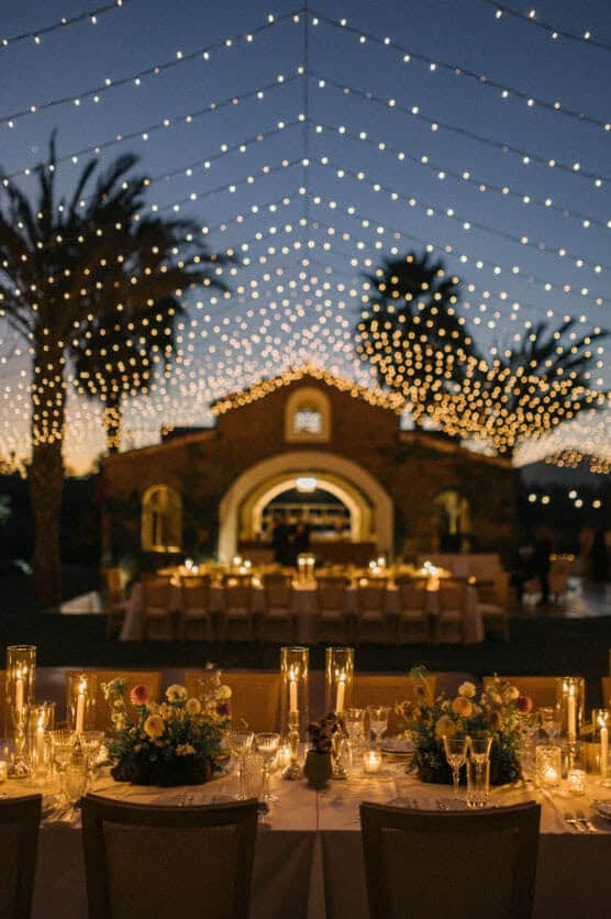 An outdoor wedding reception at dusk features long tables with floral centerpieces and candles, set under a canopy of string lights, with a building and palm trees in the background. by Justin Salem Meyer
