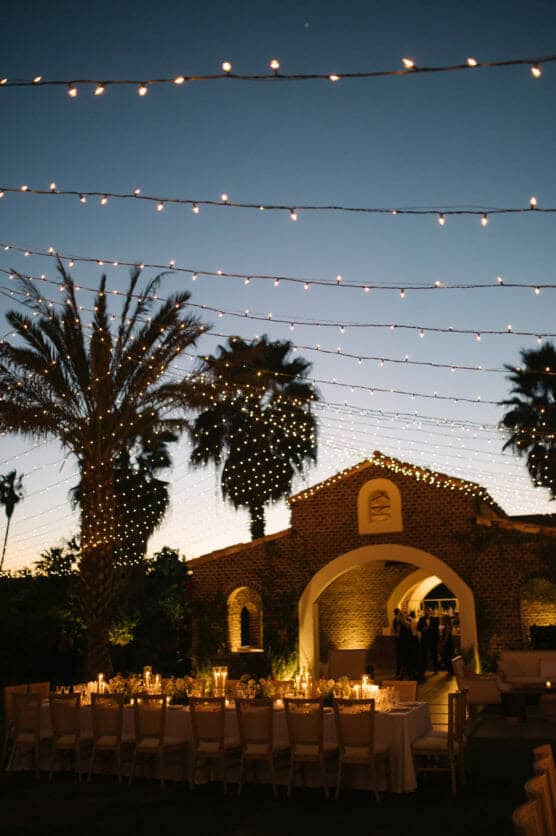A long outdoor dining table is set for an evening event under string lights, with palm trees and a brick archway building in the background at sunset. by Justin Salem Meyer