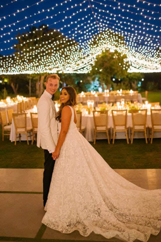 A bride in a white gown and a groom in a white jacket hold hands and smile, standing outdoors at night under a canopy of string lights, with an elegantly set dinner table behind them. by Justin Salem Meyer