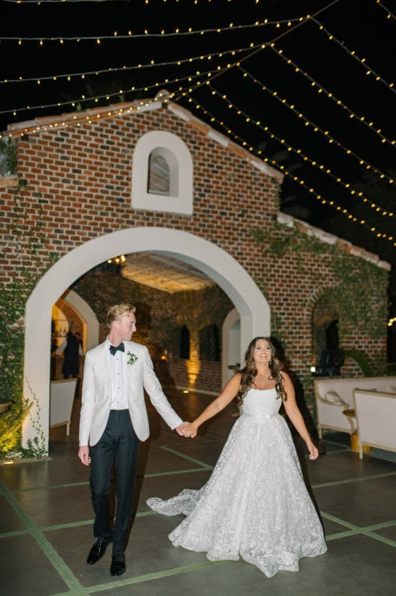 A bride in a white gown and a groom in a white tuxedo jacket hold hands and smile while walking outside under string lights, with a brick building in the background. by Justin Salem Meyer