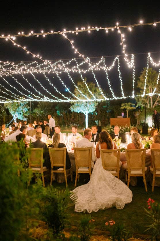 Outdoor evening wedding reception under string lights; guests in formal attire sit at a long table with candles and flowers. Bride in a white gown is visible among the guests. Trees and greenery surround the area. by Justin Salem Meyer
