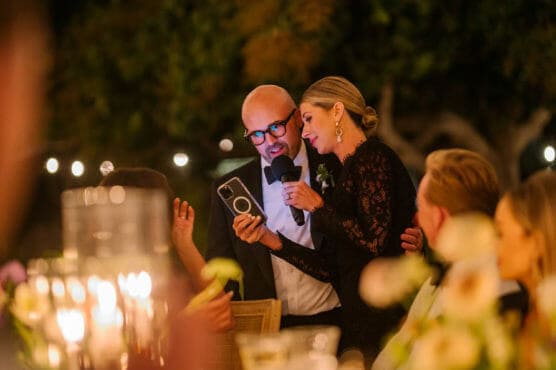 A man in a suit and a woman in a black lace dress stand close together at an outdoor event, sharing a microphone and reading from a phone, surrounded by blurred guests and candlelight. by Justin Salem Meyer