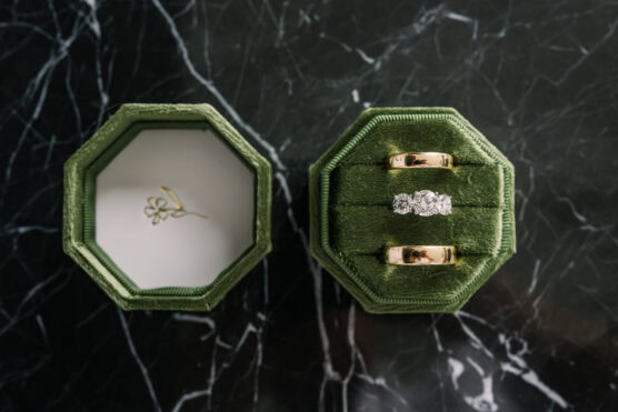 Two gold wedding bands and a diamond engagement ring rest in a green velvet ring box on a black marble surface. The open lid, lined with white fabric and a delicate floral design, sits beside the rings. by Justin Salem Meyer