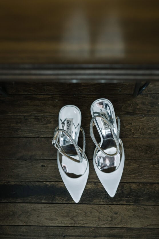A pair of white pointed-toe high-heeled shoes with silver straps are placed neatly on a wooden floor, viewed from above. by Justin Salem Meyer