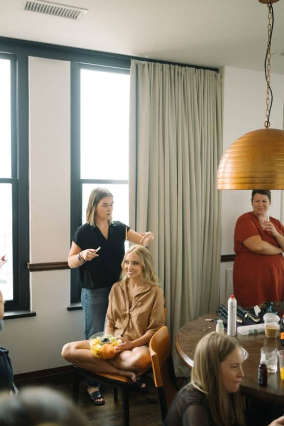 A woman sits on a chair, smiling with a fruit bowl in her lap, while another woman styles her hair. Two more women stand nearby in a bright room with large windows and a round table with hair products. by Justin Salem Meyer