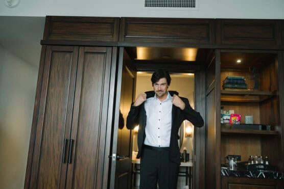 A man stands in a doorway putting on a black suit jacket over a white shirt, with wooden cabinets and shelves containing books and snacks in the background. by Justin Salem Meyer