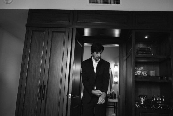 A man in a suit adjusts his sleeve while standing in front of a dark wooden wardrobe in a dimly lit room, with shelves containing various items visible to the right. by Justin Salem Meyer