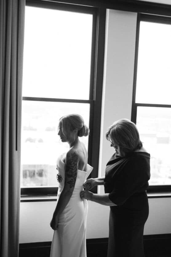 A woman helps another woman, dressed in a wedding gown, adjust her dress by a window. Soft light filters in, and the scene is in black and white, creating an intimate, emotional atmosphere. by Justin Salem Meyer