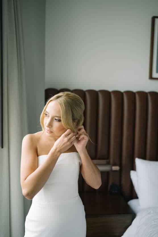 A woman in a strapless white dress stands by a window, adjusting an earring. She has blonde hair styled in an updo and is looking outside. The scene is set in a modern, softly lit bedroom. by Justin Salem Meyer