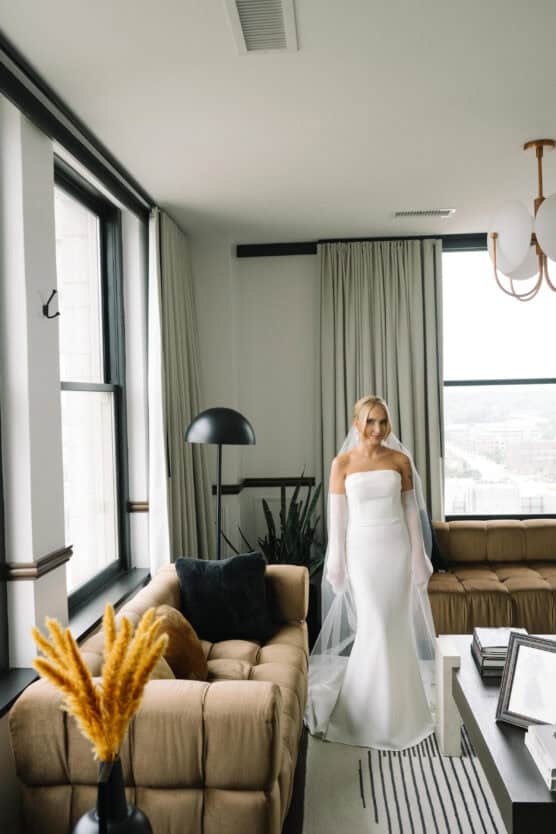 A bride in a strapless white gown and sheer gloves stands in a modern, light-filled living room with tan couches, large windows, and contemporary decor. by Justin Salem Meyer
