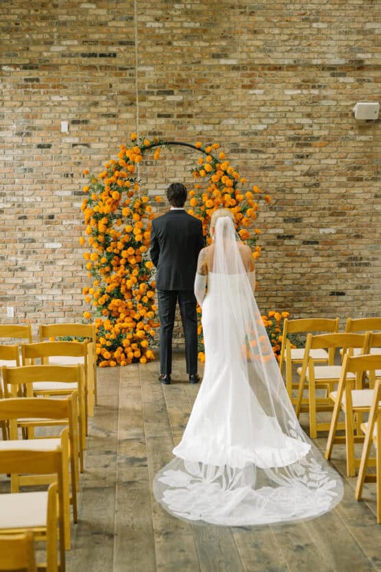 A bride in a white gown and veil walks toward a groom in a black suit, standing in front of an orange floral arch in a rustic venue with wooden chairs and a brick wall. by Justin Salem Meyer