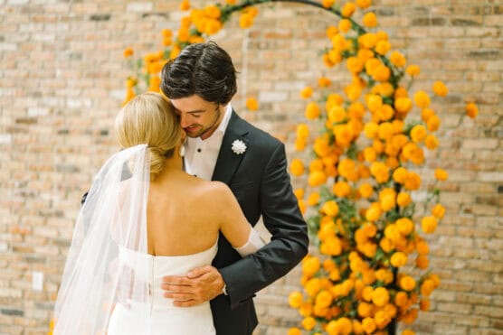 A bride and groom embrace in front of a brick wall decorated with a circular arch of vibrant yellow flowers. The bride wears a white dress and veil, and the groom wears a black tuxedo. by Justin Salem Meyer
