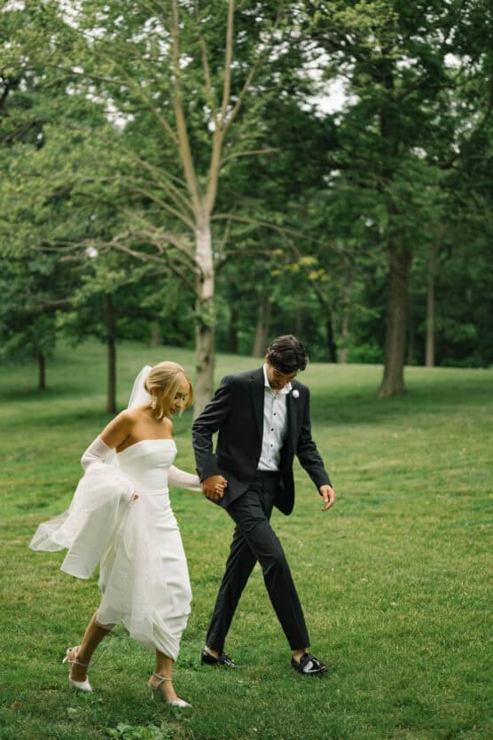 A bride in a white dress and a groom in a black suit walk hand-in-hand on green grass in a park surrounded by trees. by Justin Salem Meyer