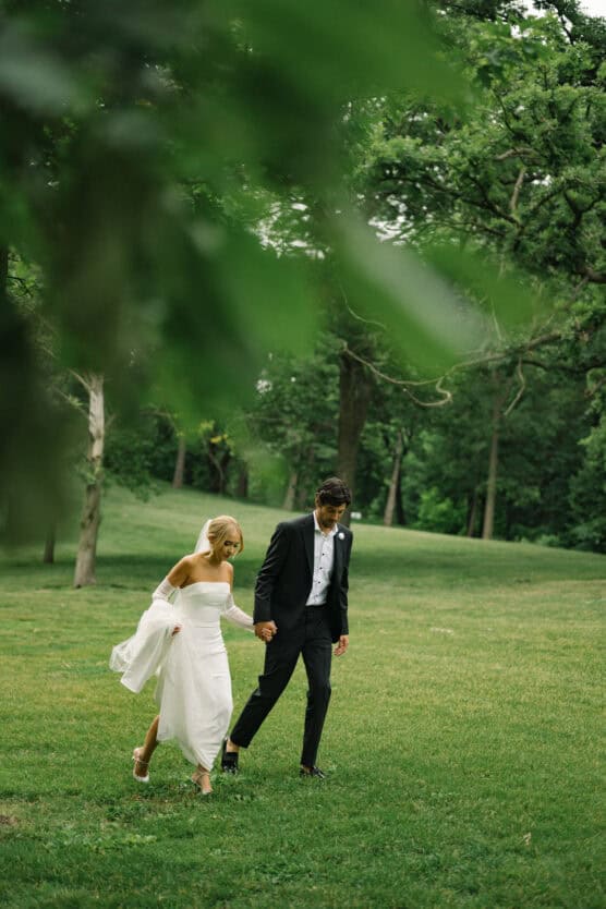 A bride and groom walk hand-in-hand across a grassy field, surrounded by lush green trees. The bride wears a strapless white dress, and the groom is in a black suit. The scene feels peaceful and romantic. by Justin Salem Meyer