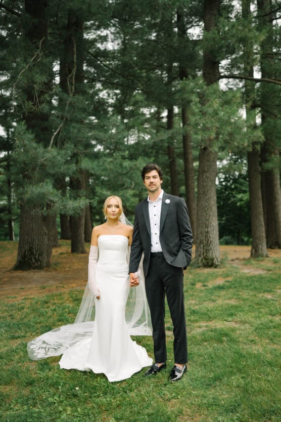 A bride in a white dress and veil stands holding hands with a groom in a dark suit and white shirt. They are outdoors in a forested area with tall pine trees and green grass. by Justin Salem Meyer