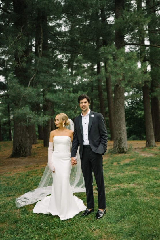 A bride in a white off-shoulder gown and veil holds hands with a groom in a dark suit and white shirt, standing together on grass among tall pine trees in a forested area. by Justin Salem Meyer